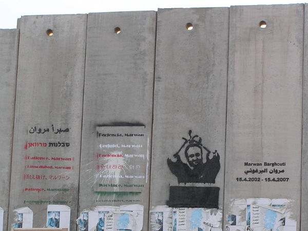 a photo of a graffiti portrait of Marwan Barghouti, bound hands above head, with text on the Israeli West Bank separation barrier, near Qalandia